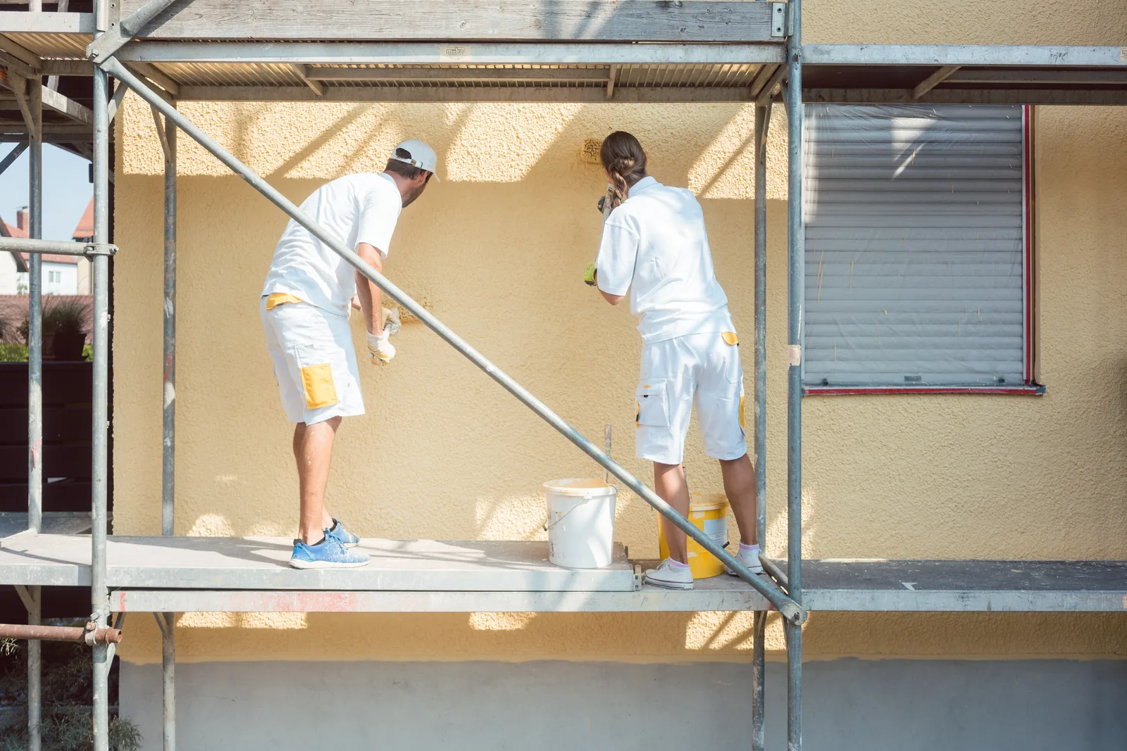 Zwei Maler tragen auf einem Gerüst Putz auf eine gelbe Hausfassade auf