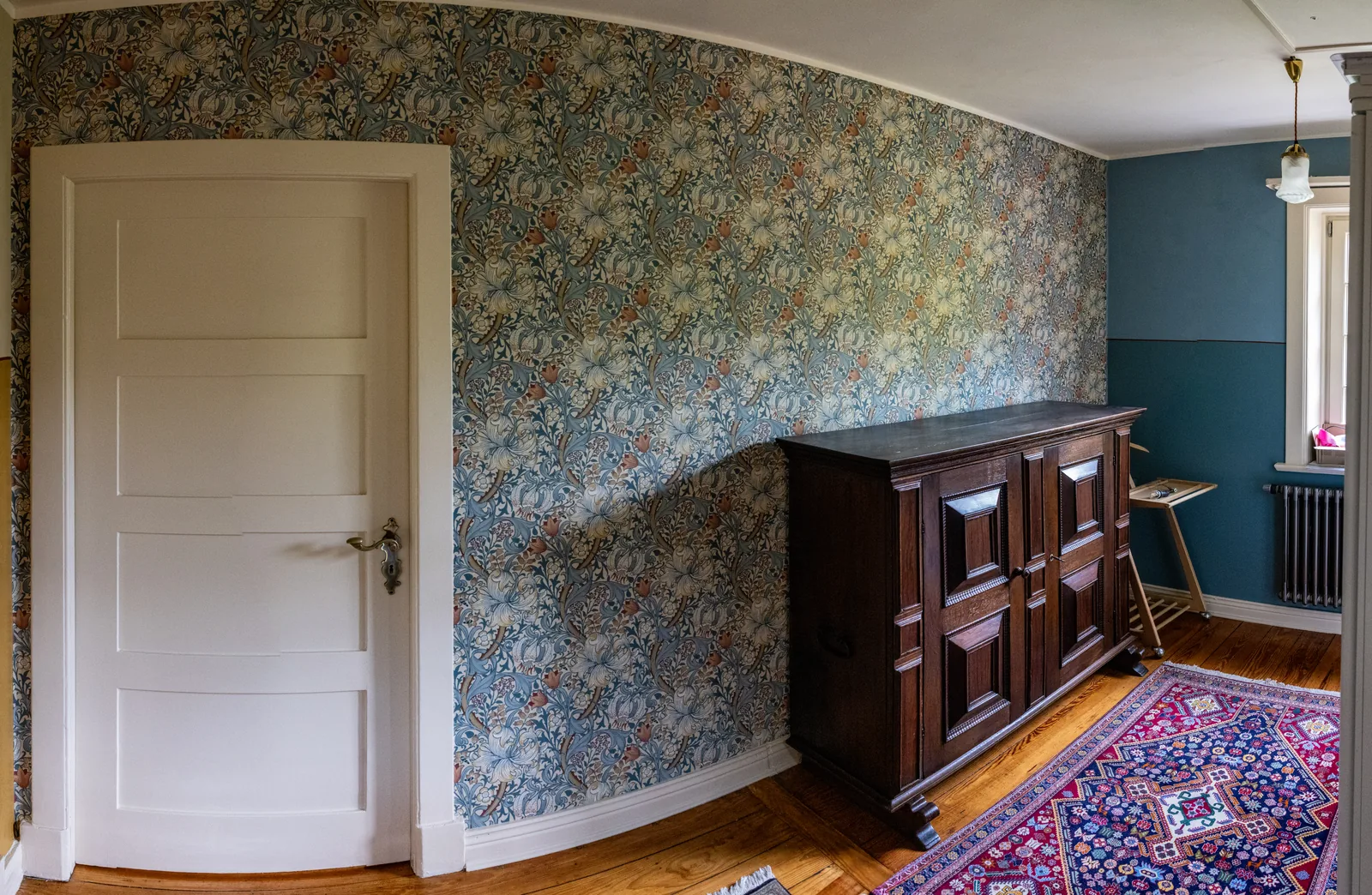 Altbauzimmer mit großflächiger Blumentapete, weißer Flügeltür und alter Holzkommode